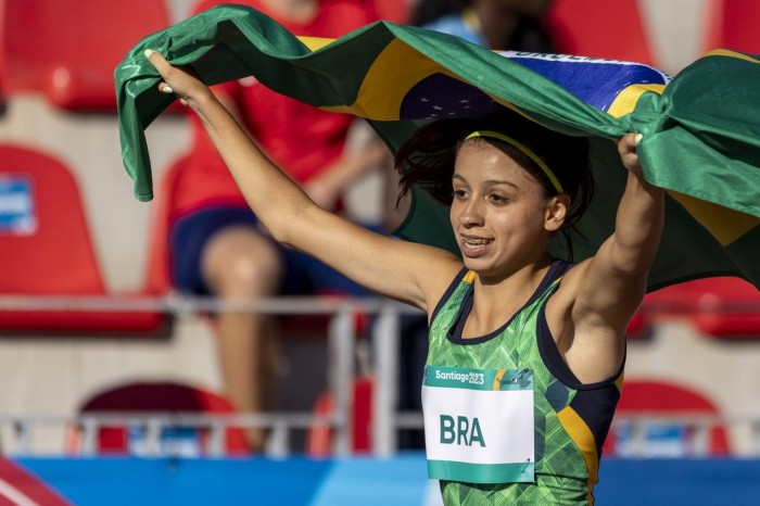 Marcelly na pista de atletismo com os braços levantados segurando bandeira. (Foto: Alessandra Cabral/CPB)